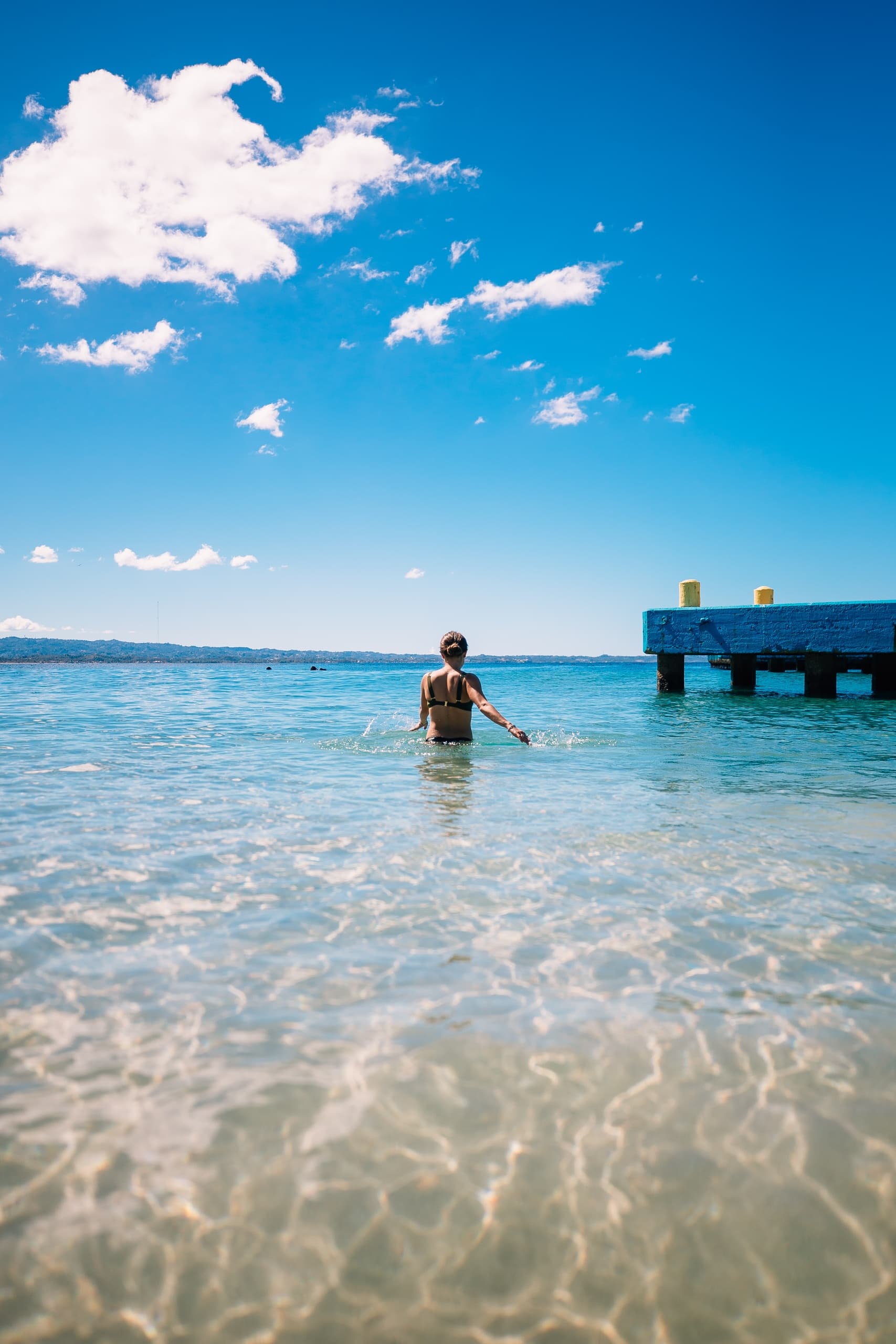 Person wading in clear tropical bay near wooden pier at noon