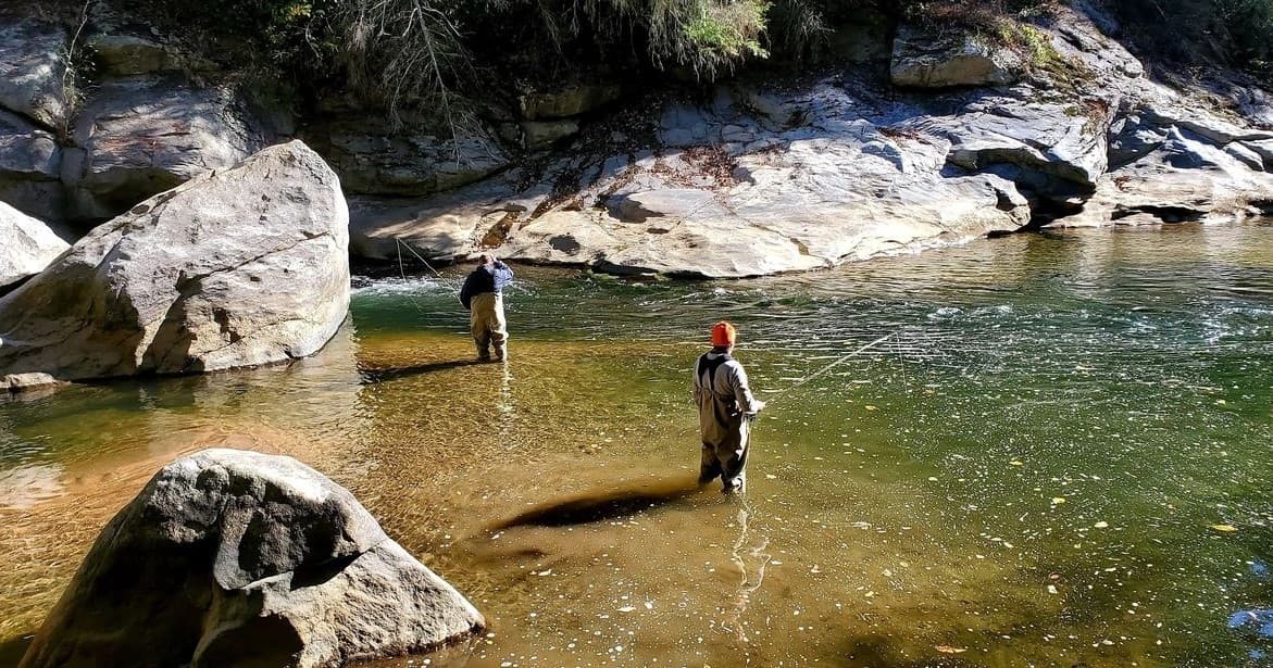 Two anglers fly fishing in shallow rocky river, sunlit scene