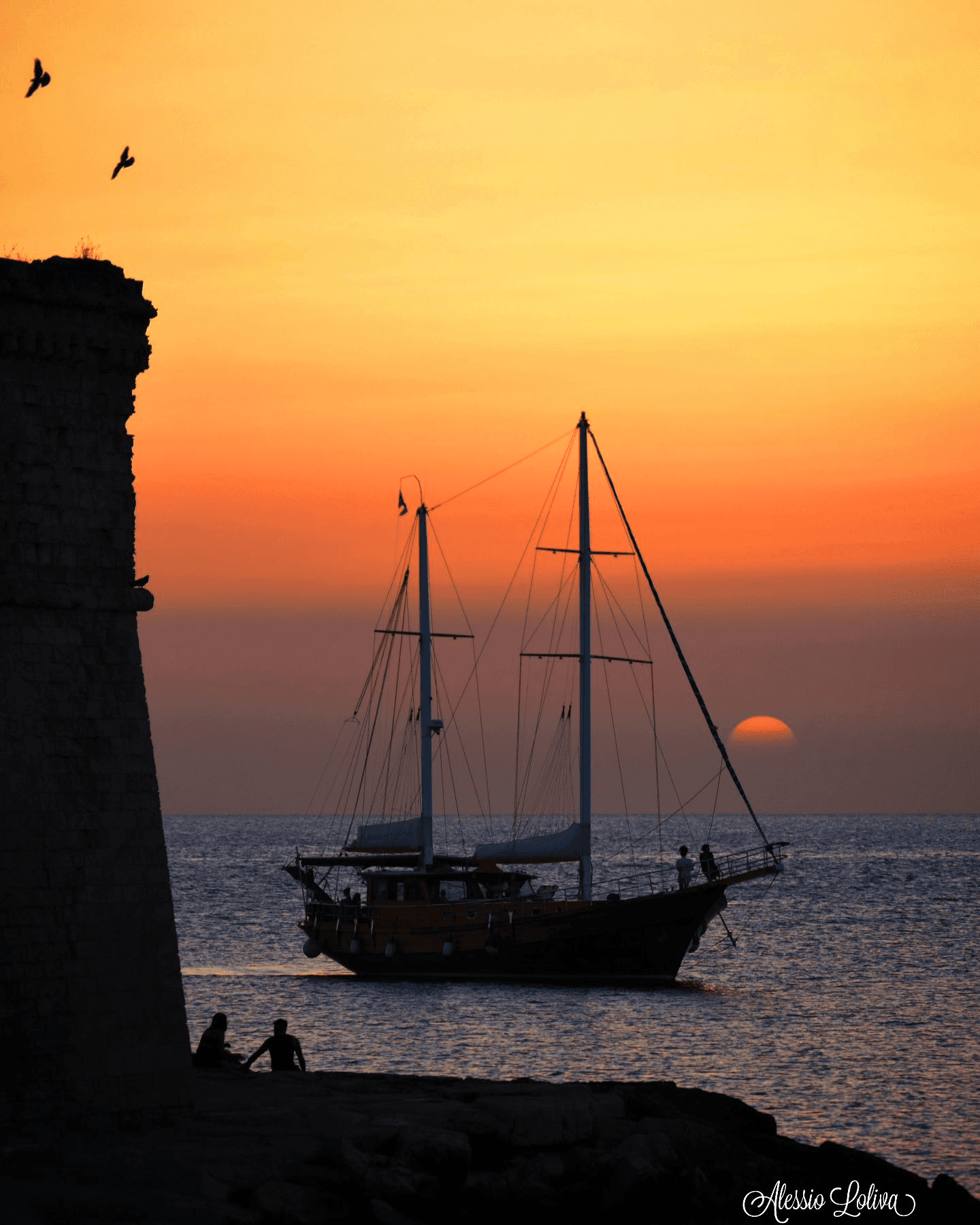 Sailboat silhouetted at sunset off rocky Puglia coastline