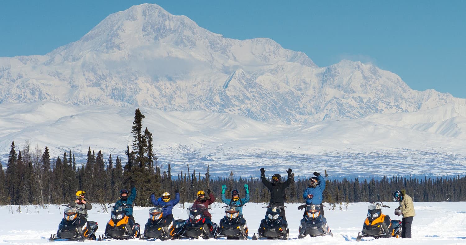 Snowmobilers riding across snowy plain with Denali mountain backdrop