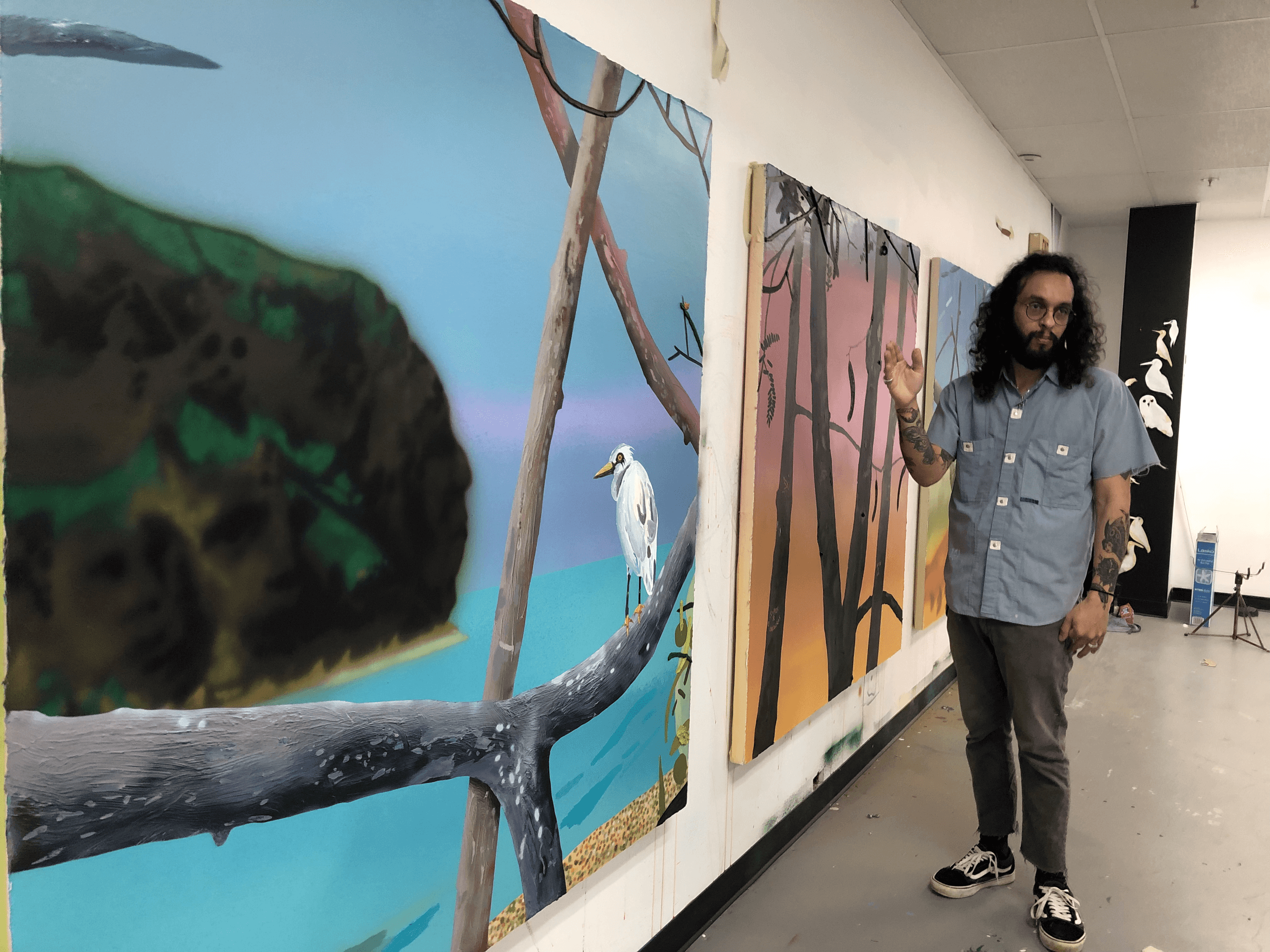 Artist standing beside large nature murals in indoor gallery
