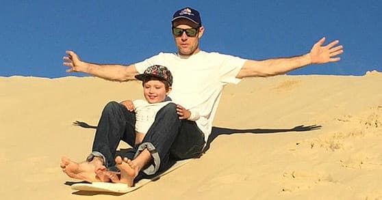 Father and child sandboarding together down smooth dune slope
