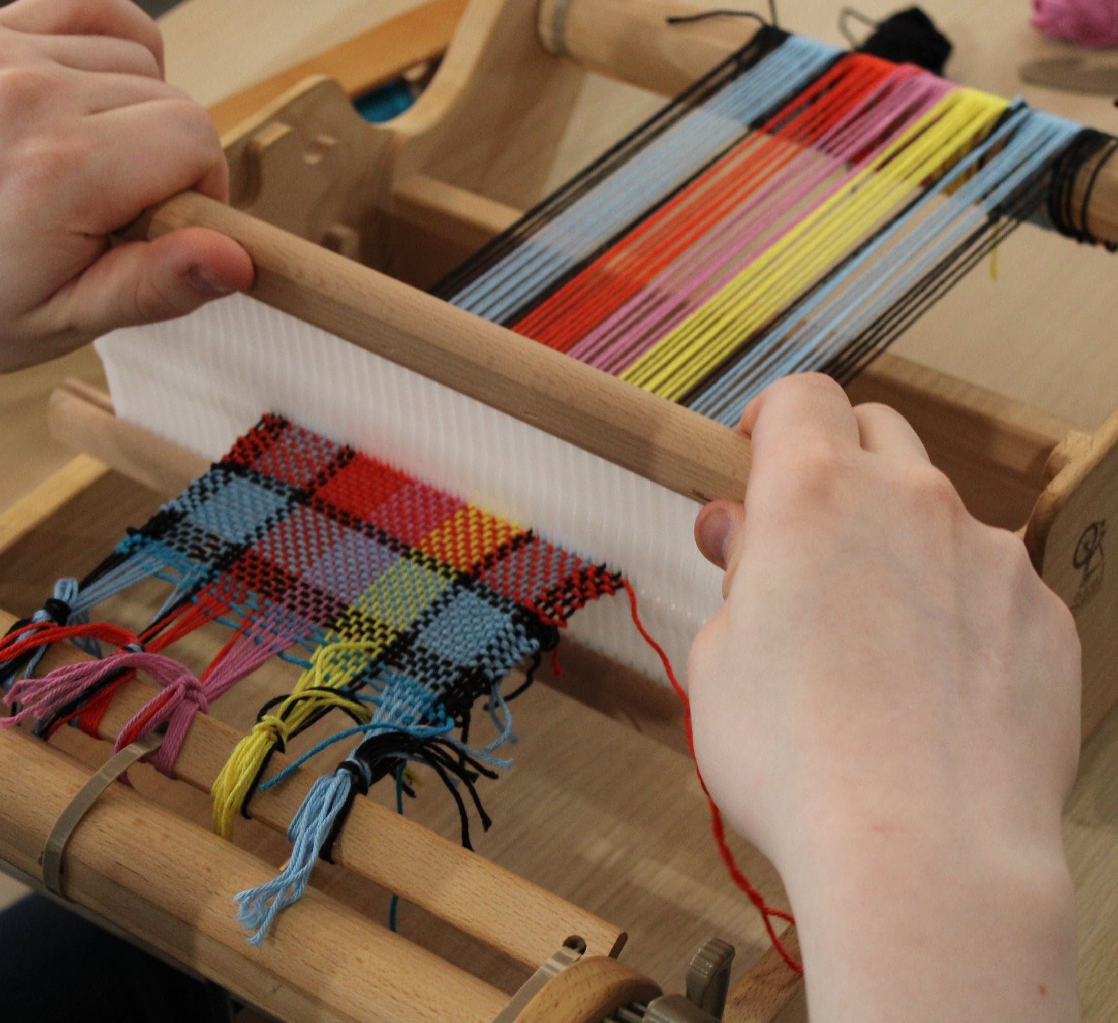 Hands weaving colorful plaid fabric on a small indoor loom