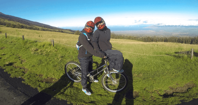 Two riders sharing a mountain bike on grassy slope with volcanic views