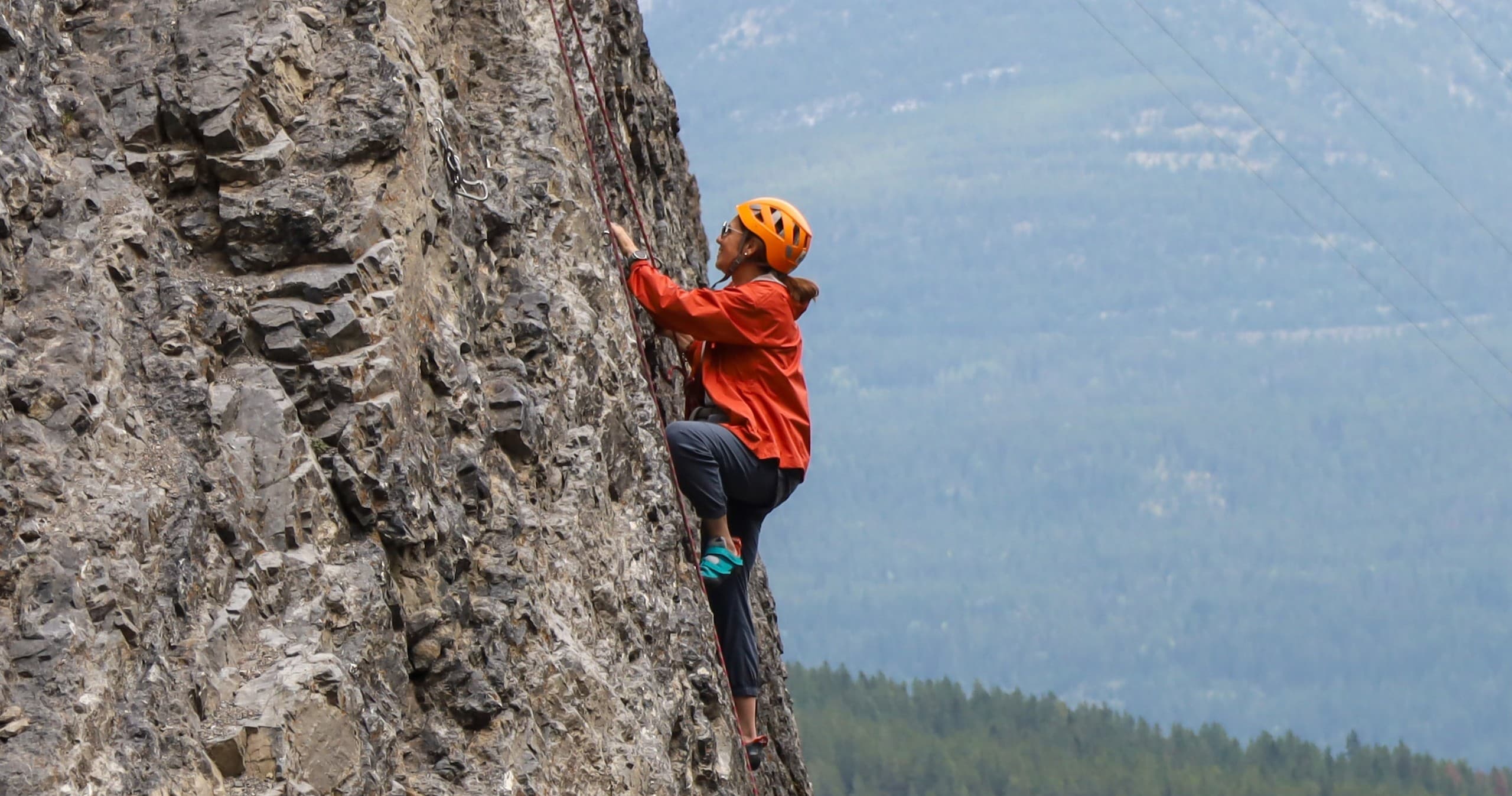 Rock climber scaling steep cliff face with forested mountain backdrop