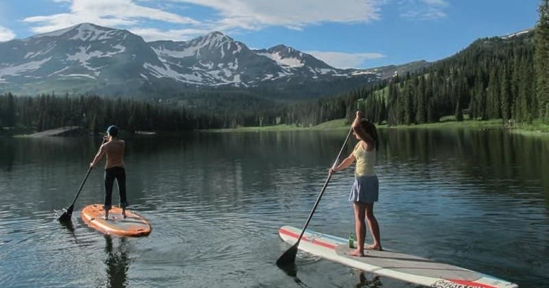 Two paddleboarders on calm mountain lake with snowy peaks