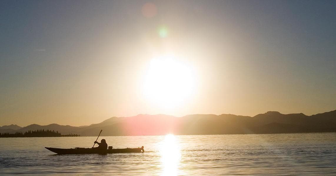 Kayaker silhouetted paddling at sunset over calm alpine lake