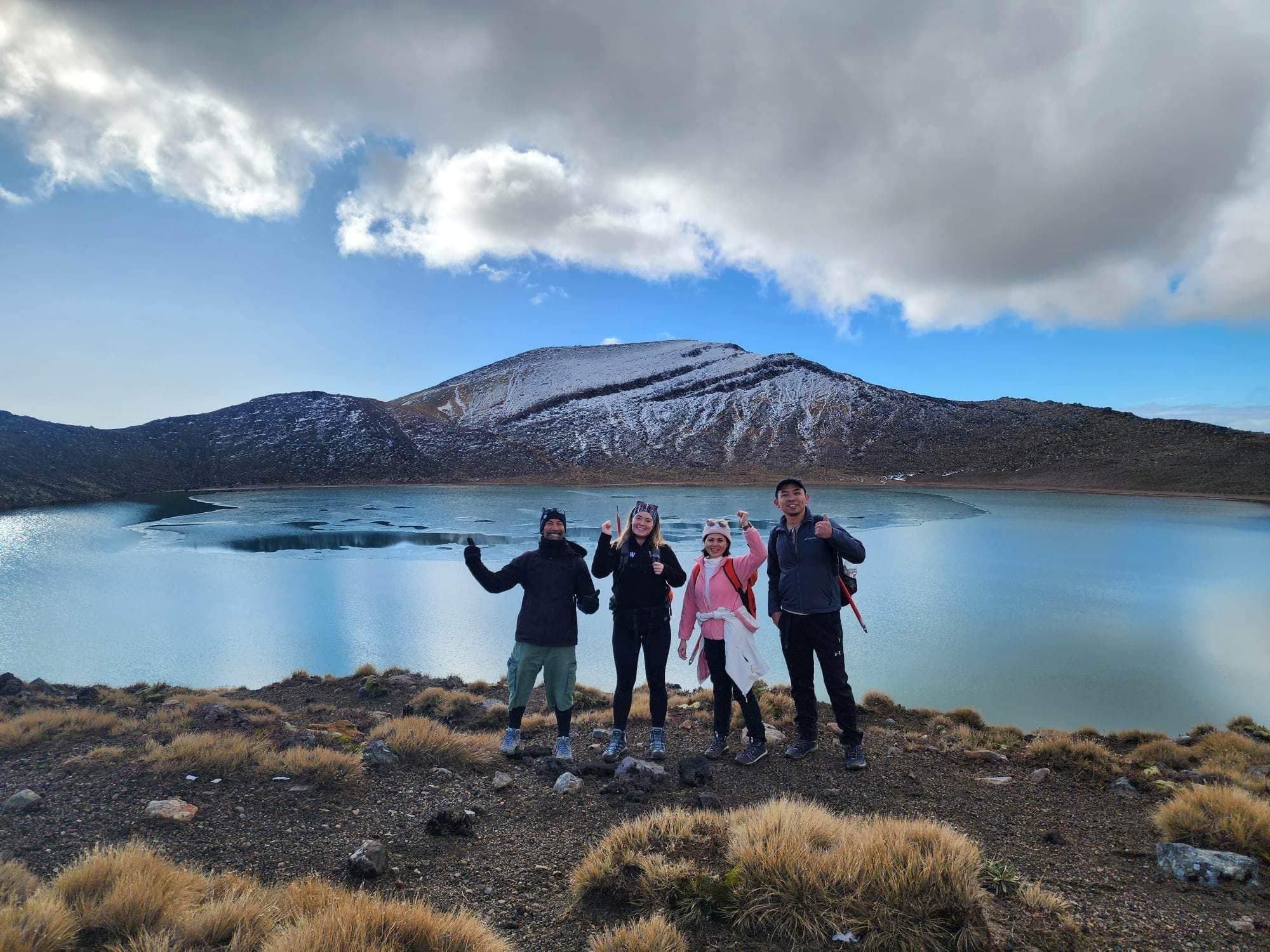 Group of hikers posing at crater lake shore with snowy peak