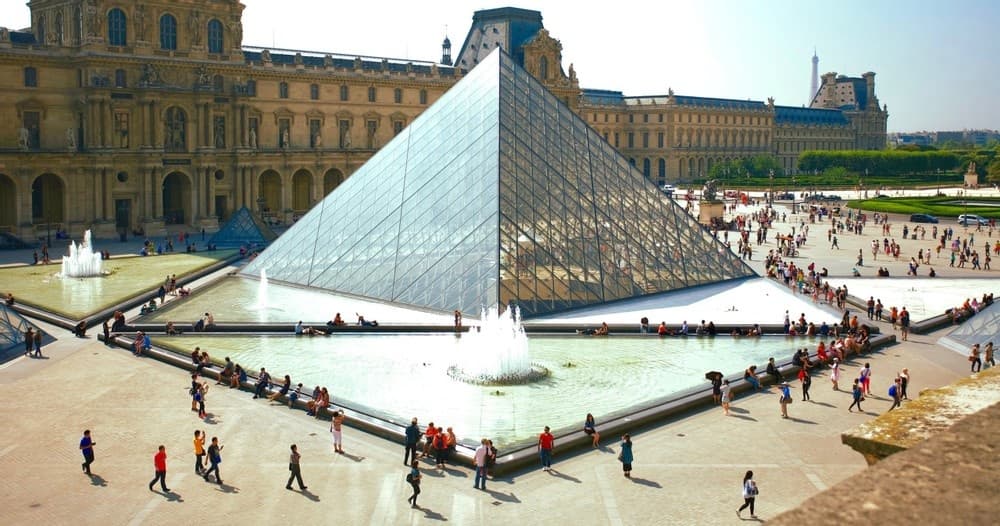 Visitors exploring Louvre Pyramid courtyard in Paris, sunny day