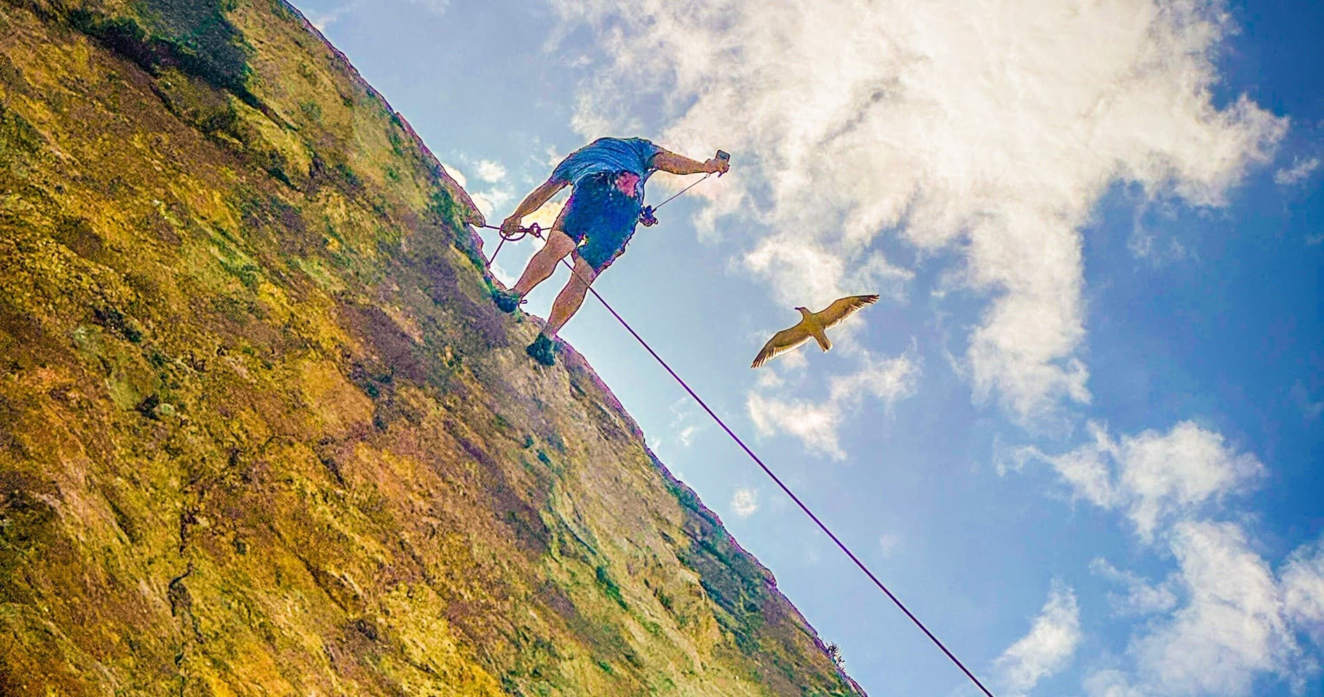 Solo climber scaling red cliff above pebble beach under blue sky