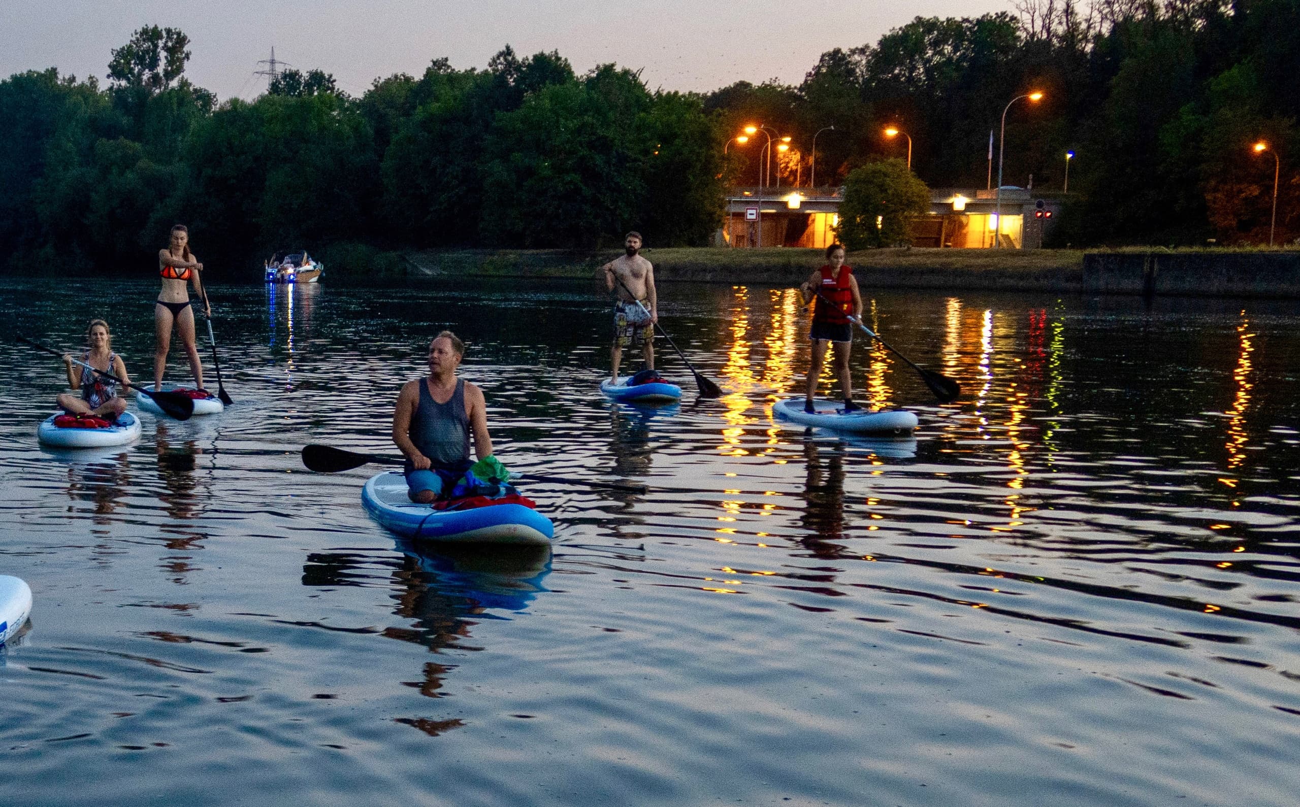 Sunset paddleboarders on a calm river with shoreline lights