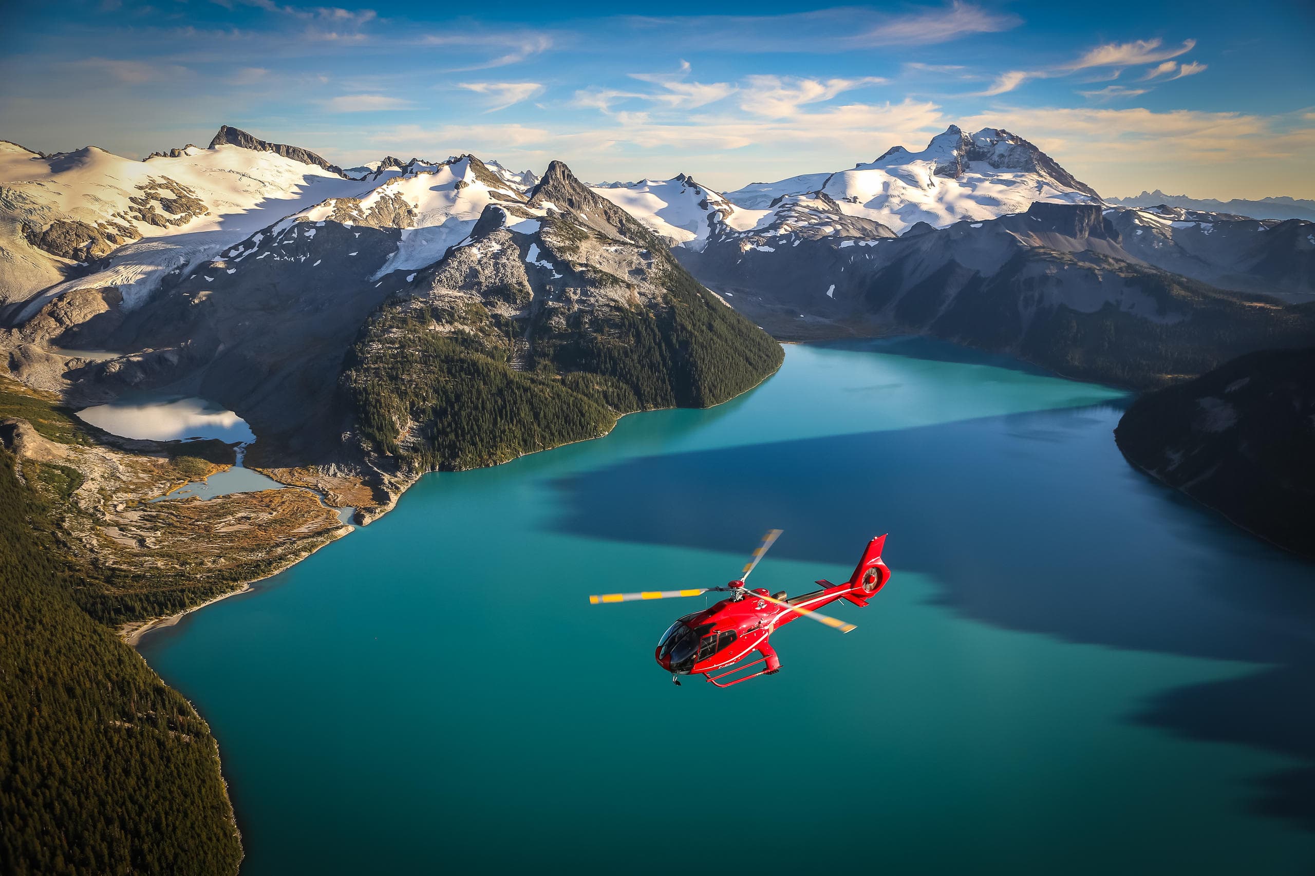 Red helicopter flying above turquoise alpine lake and rugged mountains