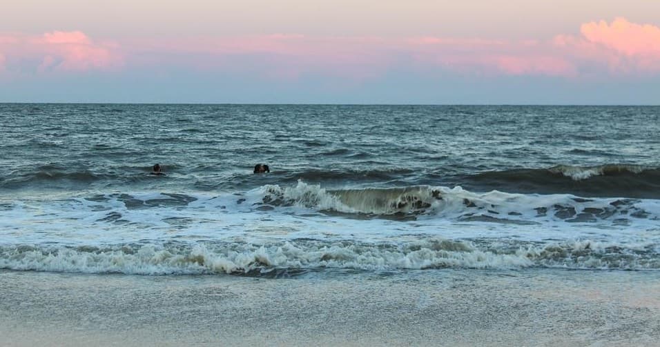 Waves breaking on sandy beach with surfers at sunset shore