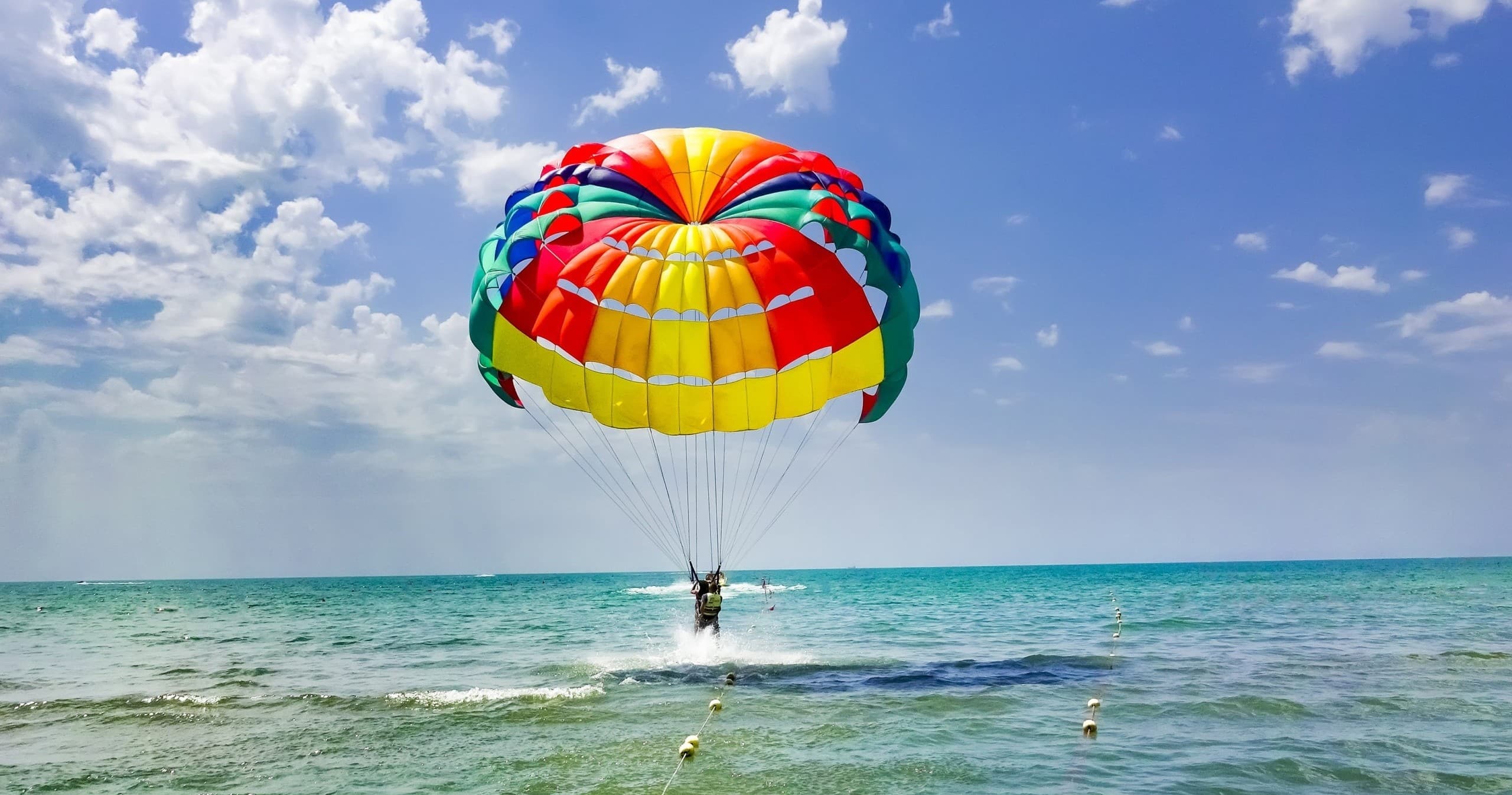 Parasailing over turquoise ocean near a sunny beach, rider splashing