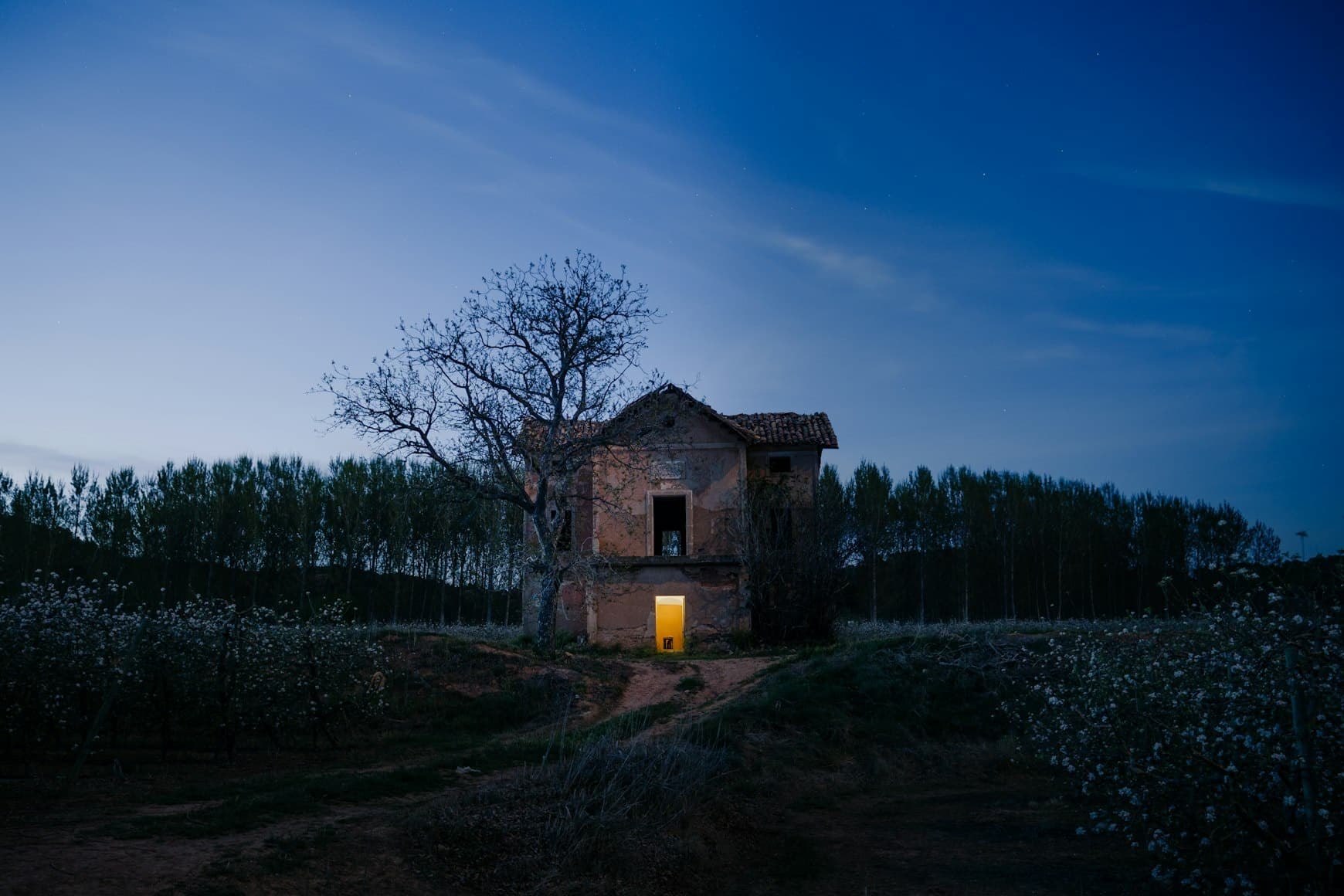 Abandoned farmhouse at dusk in rural field with illuminated door