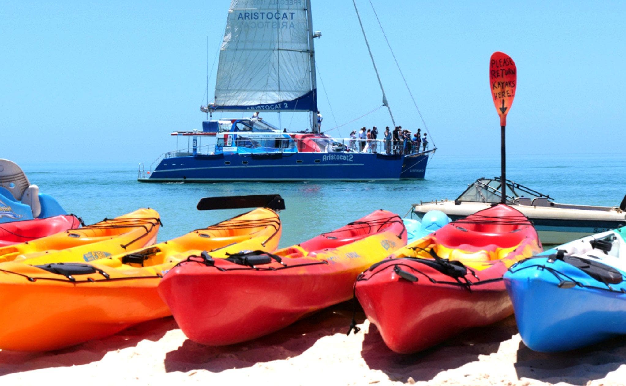 Colorful kayaks on white sand with catamaran in background