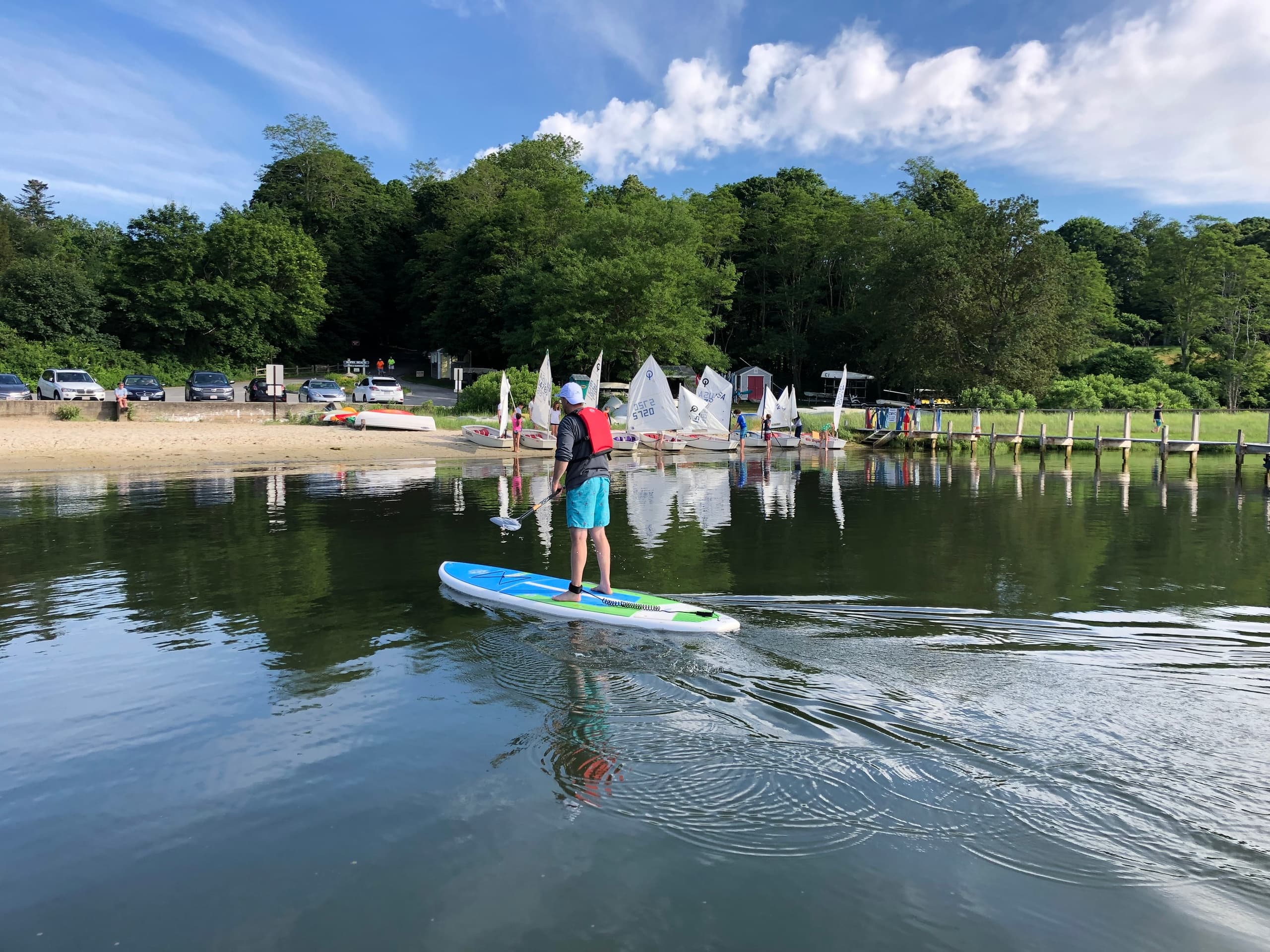 Stand-up paddleboarder gliding on calm bay near sailboats and wooded shore