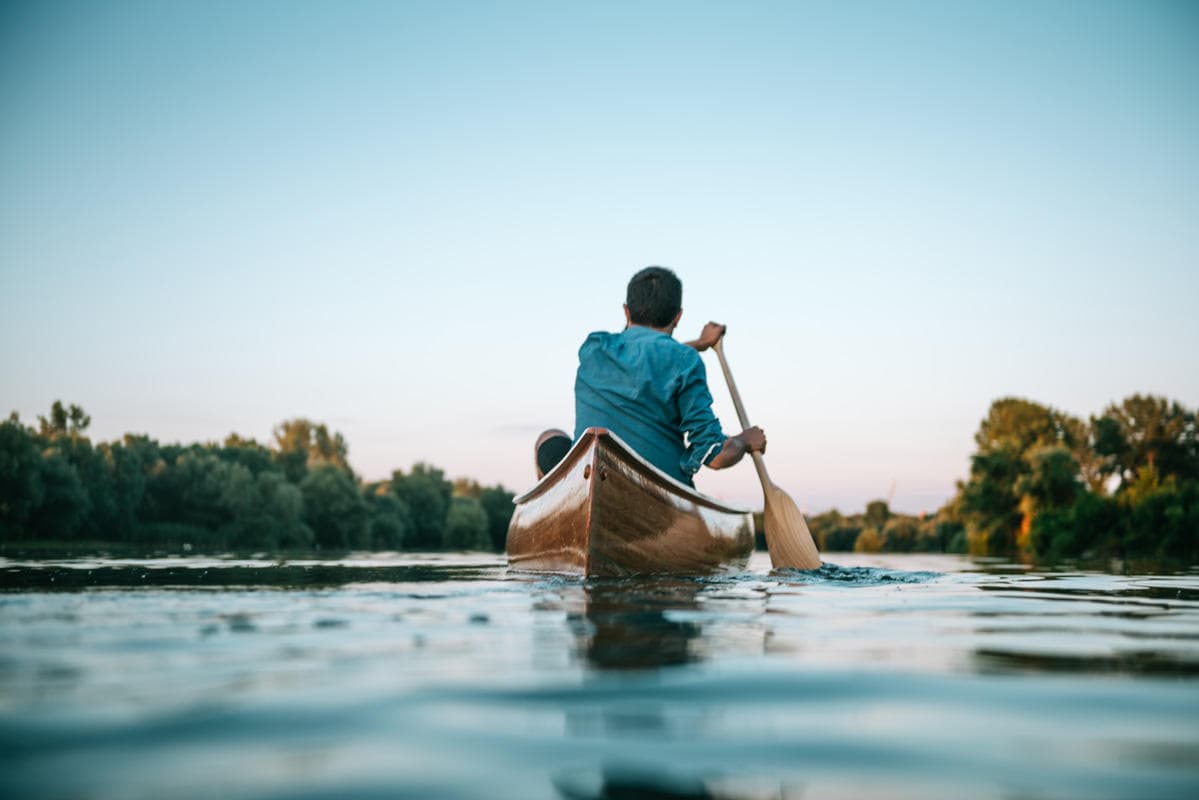 Canoeist paddling on calm river at sunrise with forested banks