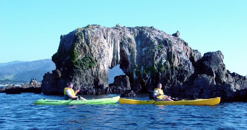 Two kayakers paddling by sea rock arch off rugged coastline