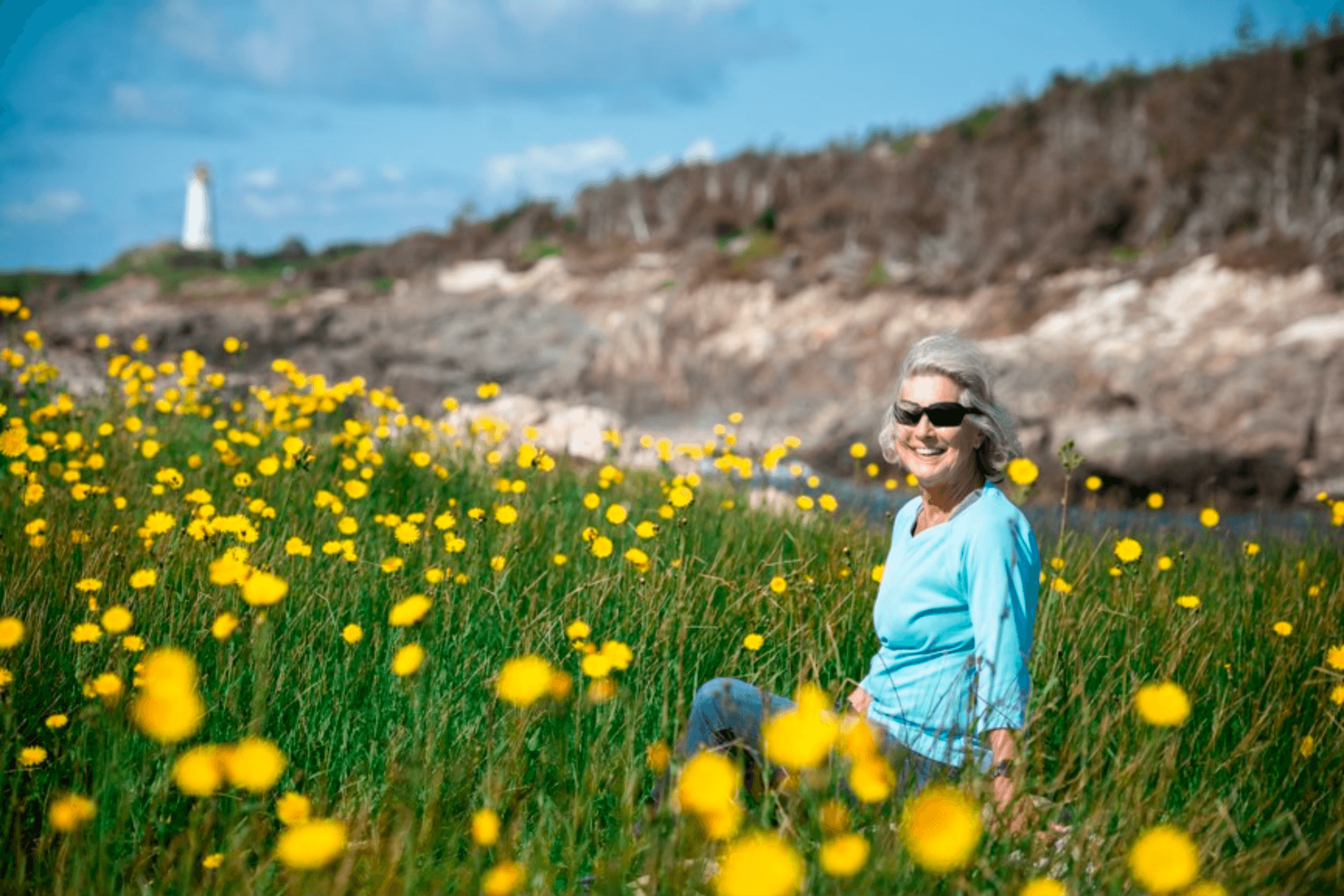 Woman sitting in coastal wildflower field near lighthouse, sunny