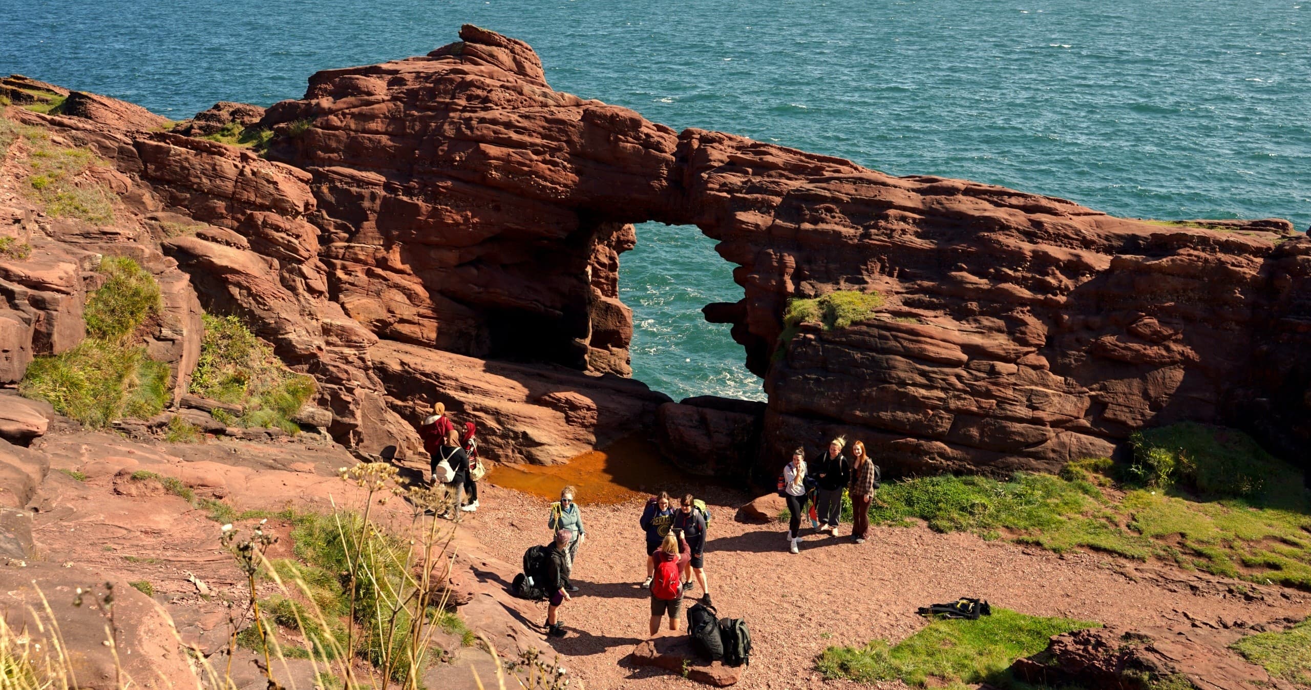 Hikers exploring coastal red sandstone arch at seaside cliffs