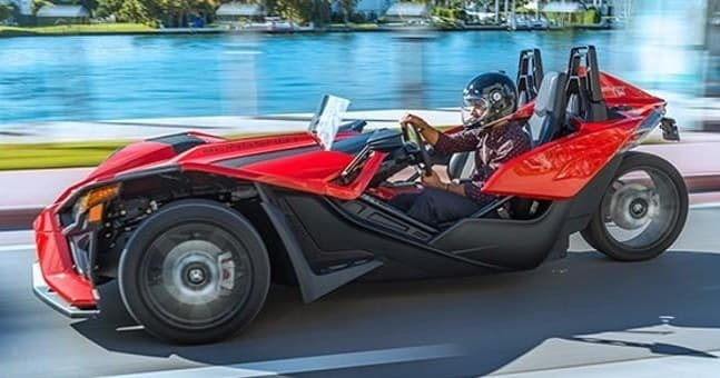 Red Polaris Slingshot driving along waterfront road with driver