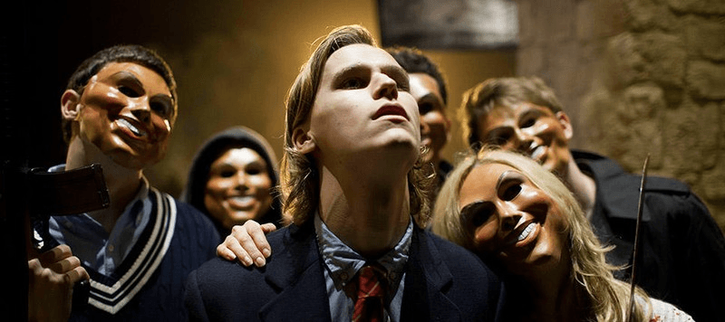 Group of masked youths surrounding central teen in dim stone interior