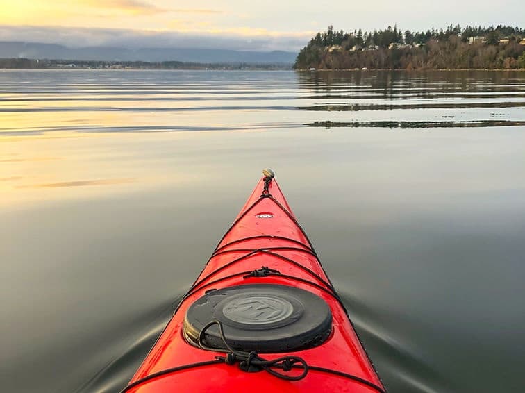Red kayak bow cutting calm reflective water at sunrise