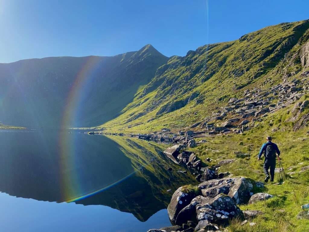 Hiker walking beside reflective mountain lake with rainbow