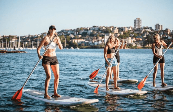Women standup paddleboarding in calm bay with city shoreline
