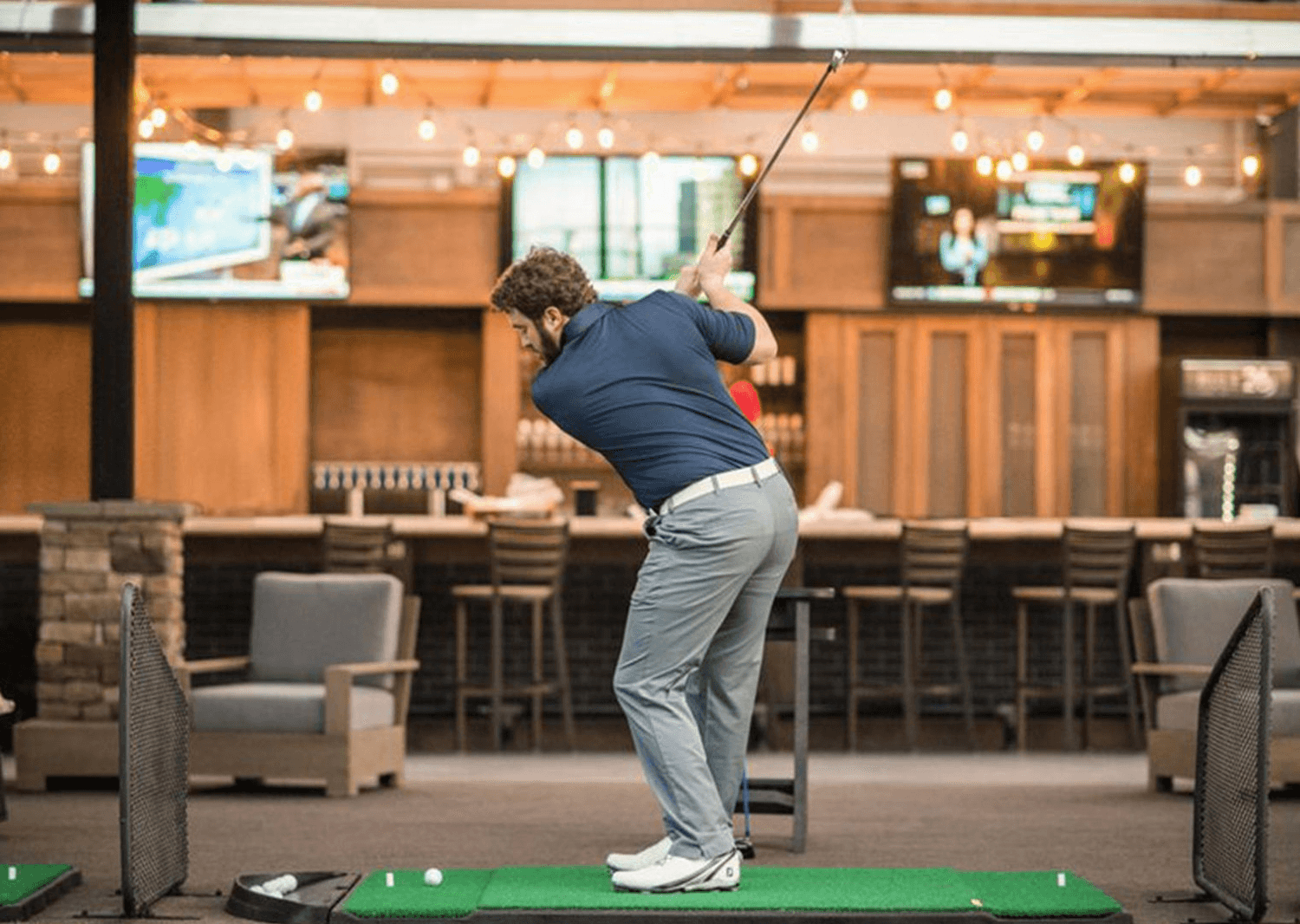 Golfer swinging at indoor driving range bay with bar seating