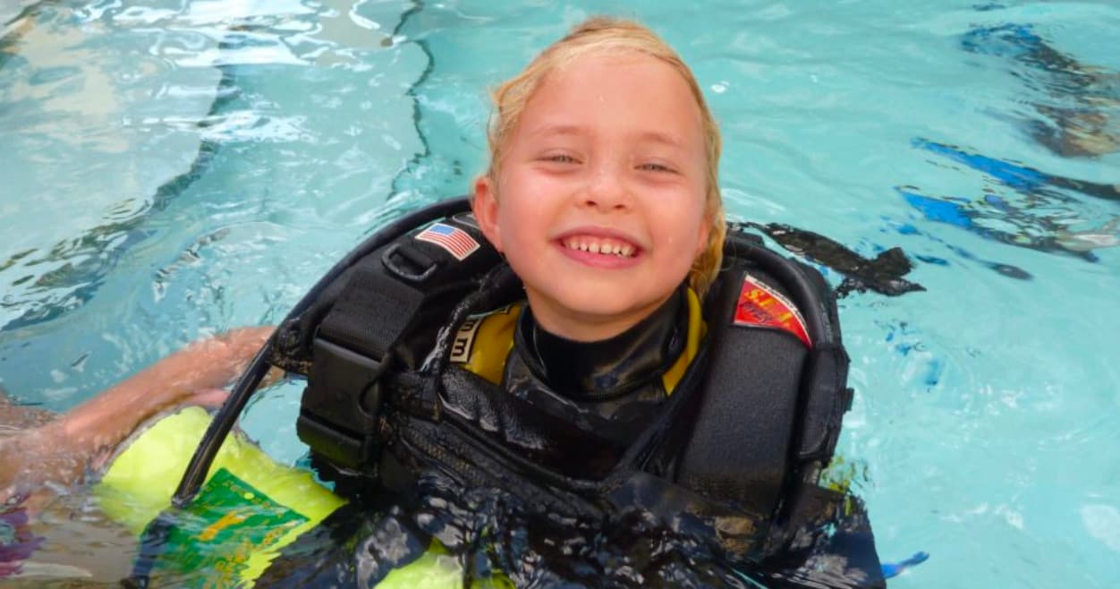 Child learning scuba in shallow training pool smiling with gear