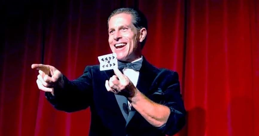 Magician performing card trick on stage with red curtain