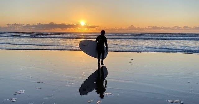 Surfer carrying board on reflective beach at sunset in UK