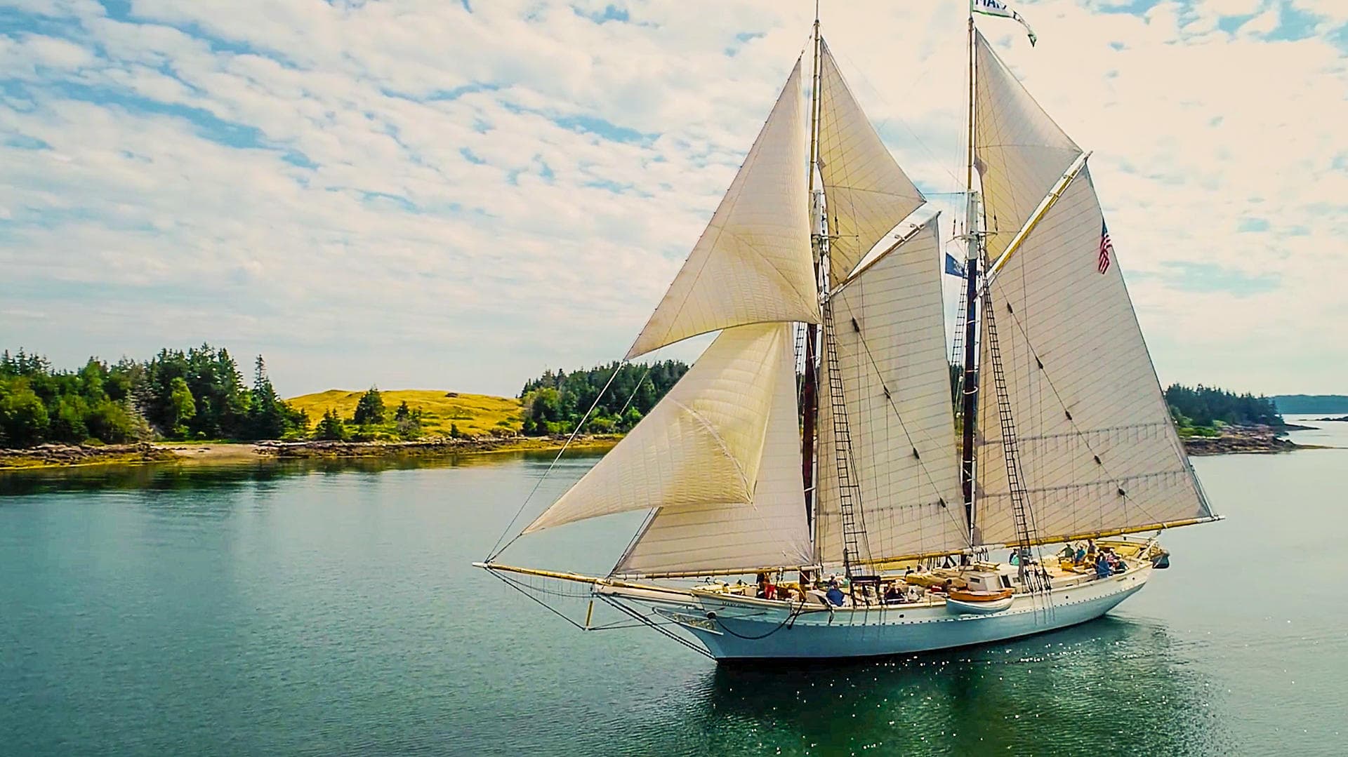 Traditional schooner sailing past tree-lined island in calm bay