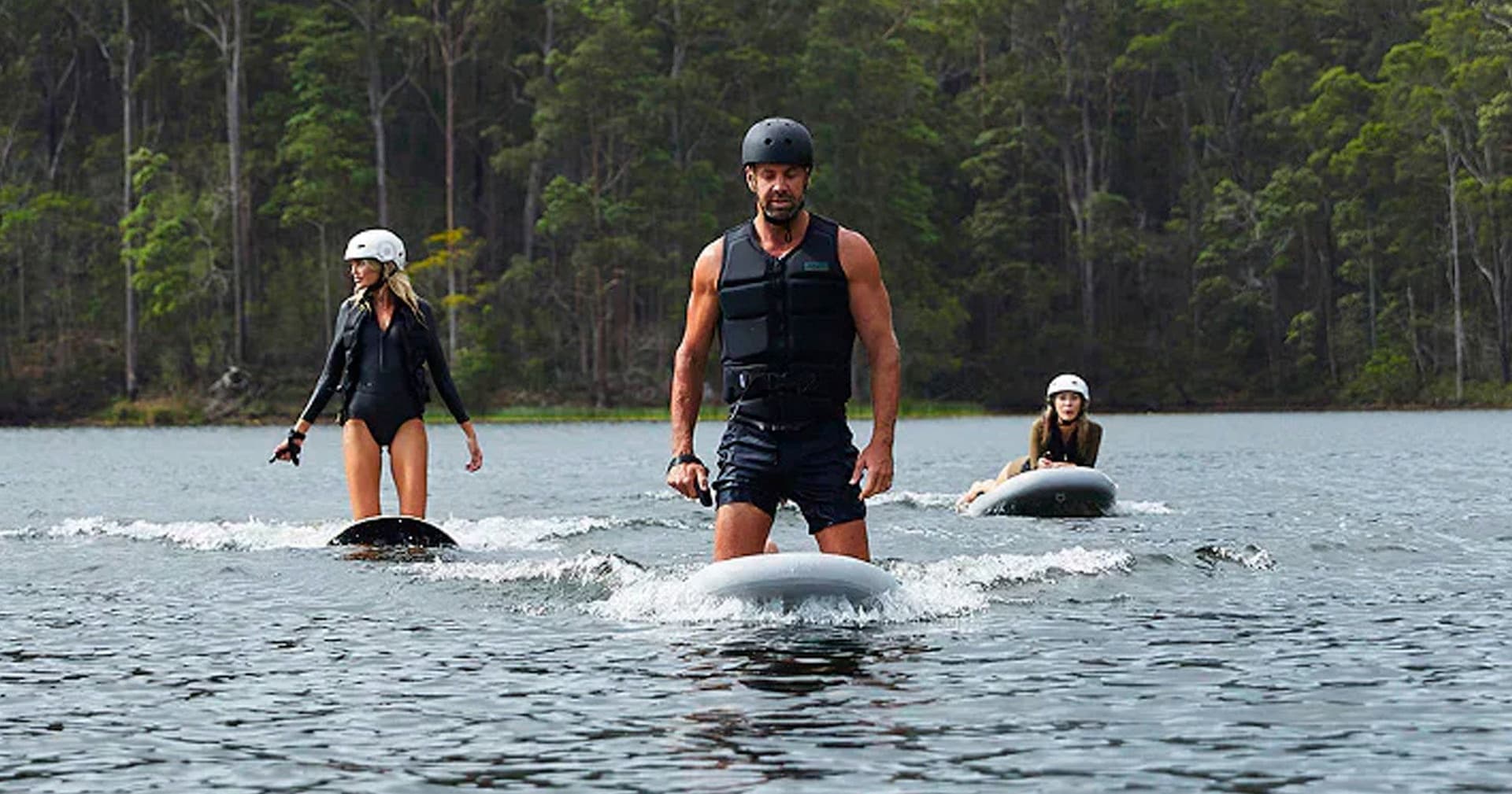 Group riding electric foil boards on lake near forested shore