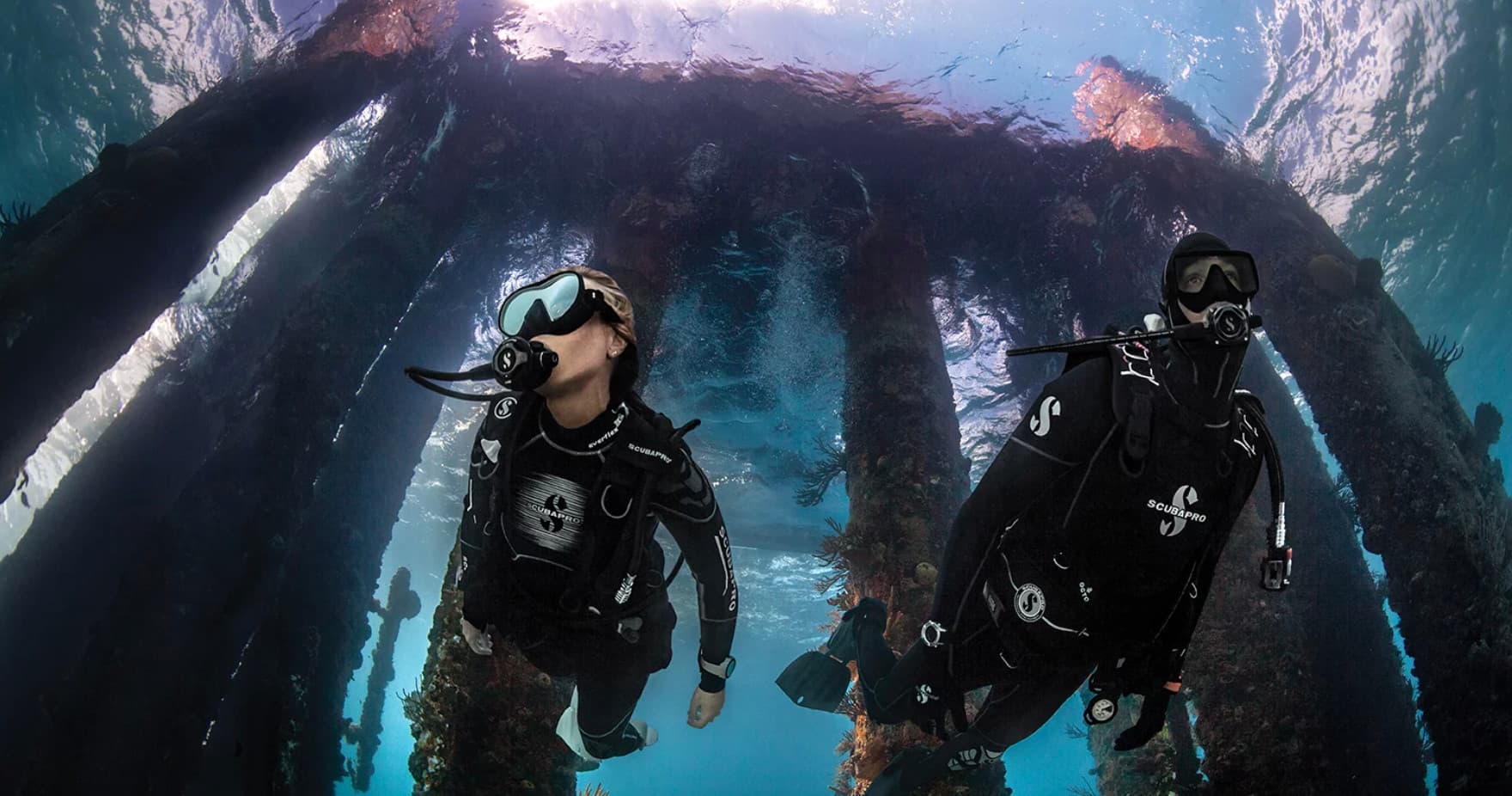 Scuba divers exploring sunlit pier pillars in clear ocean water