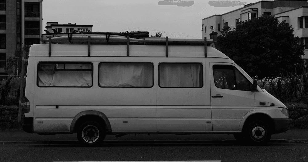 Long shot of white shuttle van parked by urban greenery, street