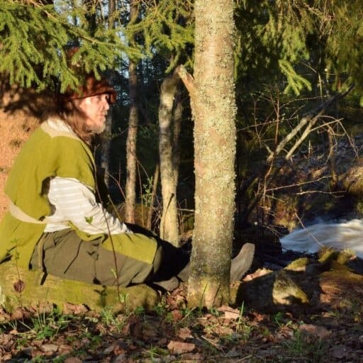 Solo adventurer resting against tree by forest stream in sunlight