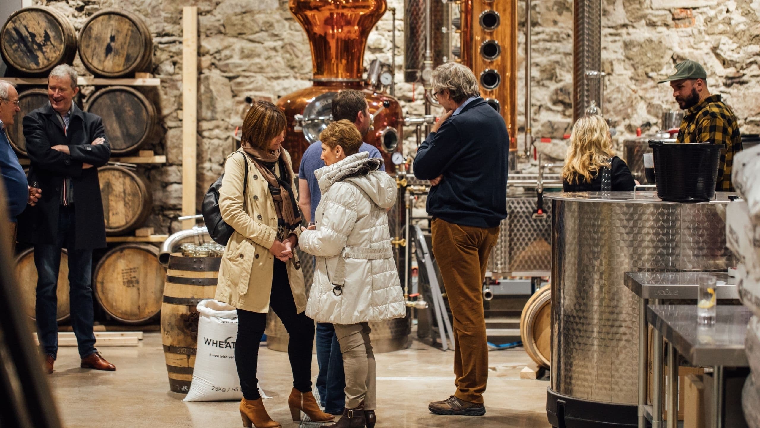 Visitors tasting and touring craft distillery floor with barrels
