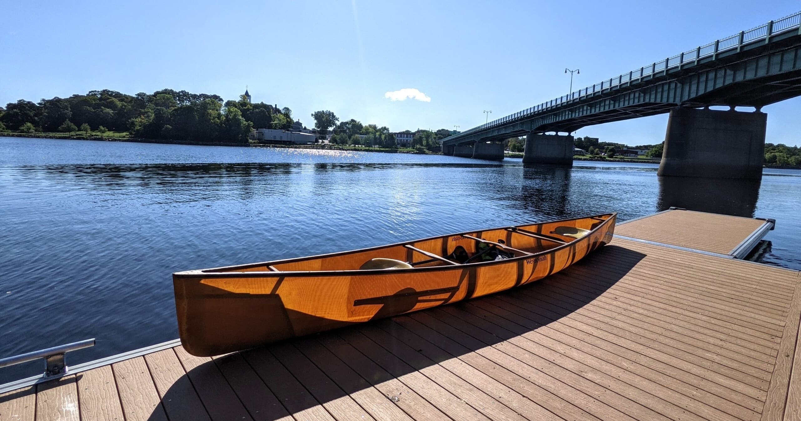 Wooden canoe moored on dock by river with bridge backdrop