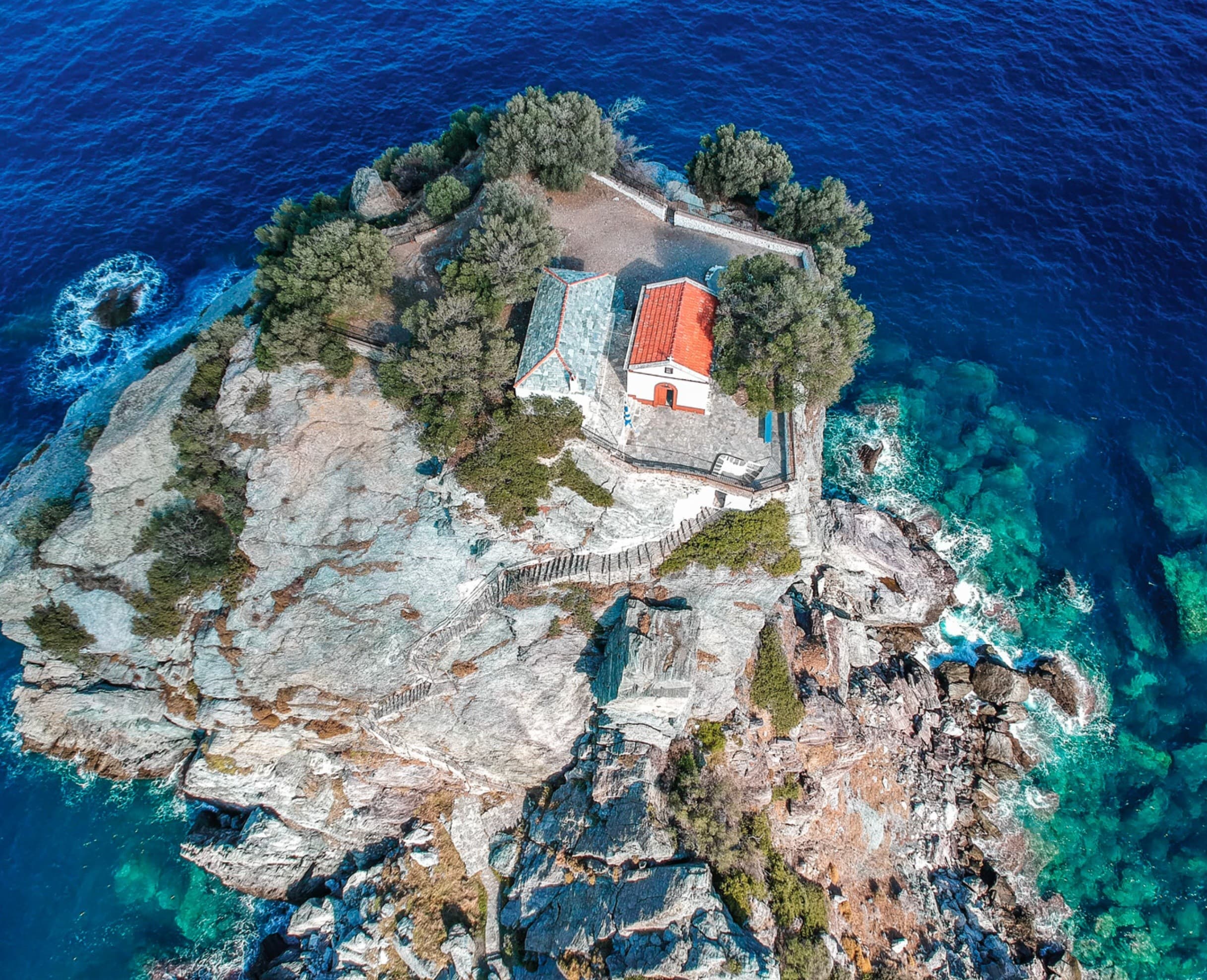 Aerial islet with red-roof house on rocky coast, scenic boat tour