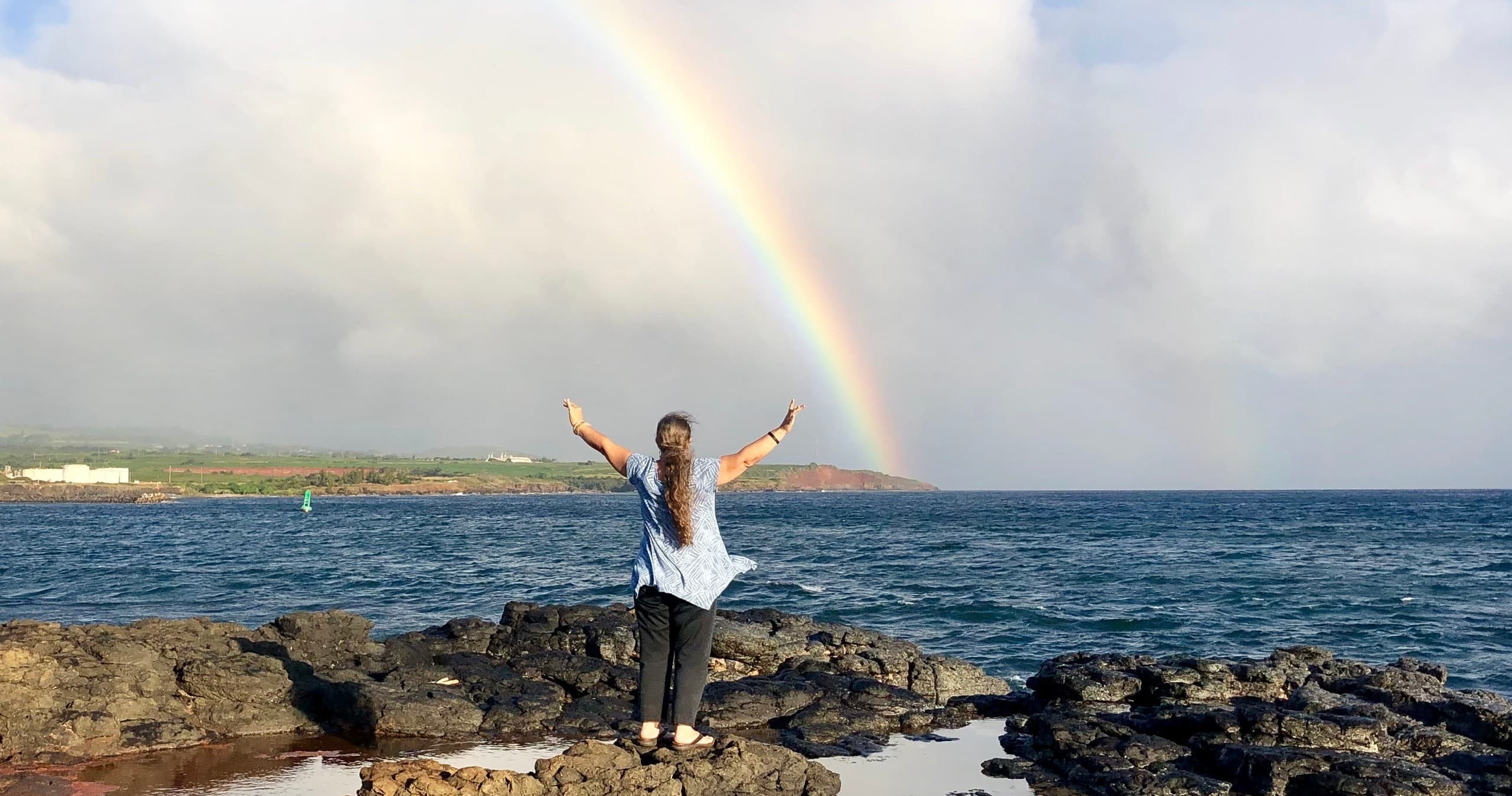 Traveler standing on rocky Kauai shore under vibrant rainbow