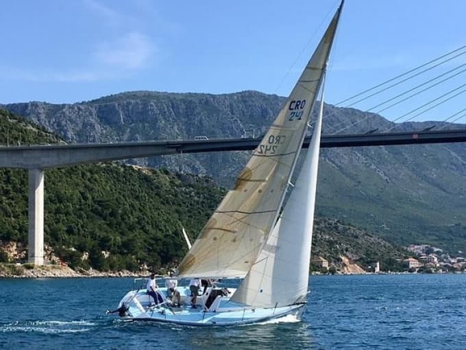 Racing sailboat under coastal bridge with mountainous backdrop