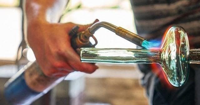 Glassblower blowing elongated vessel at bench in studio