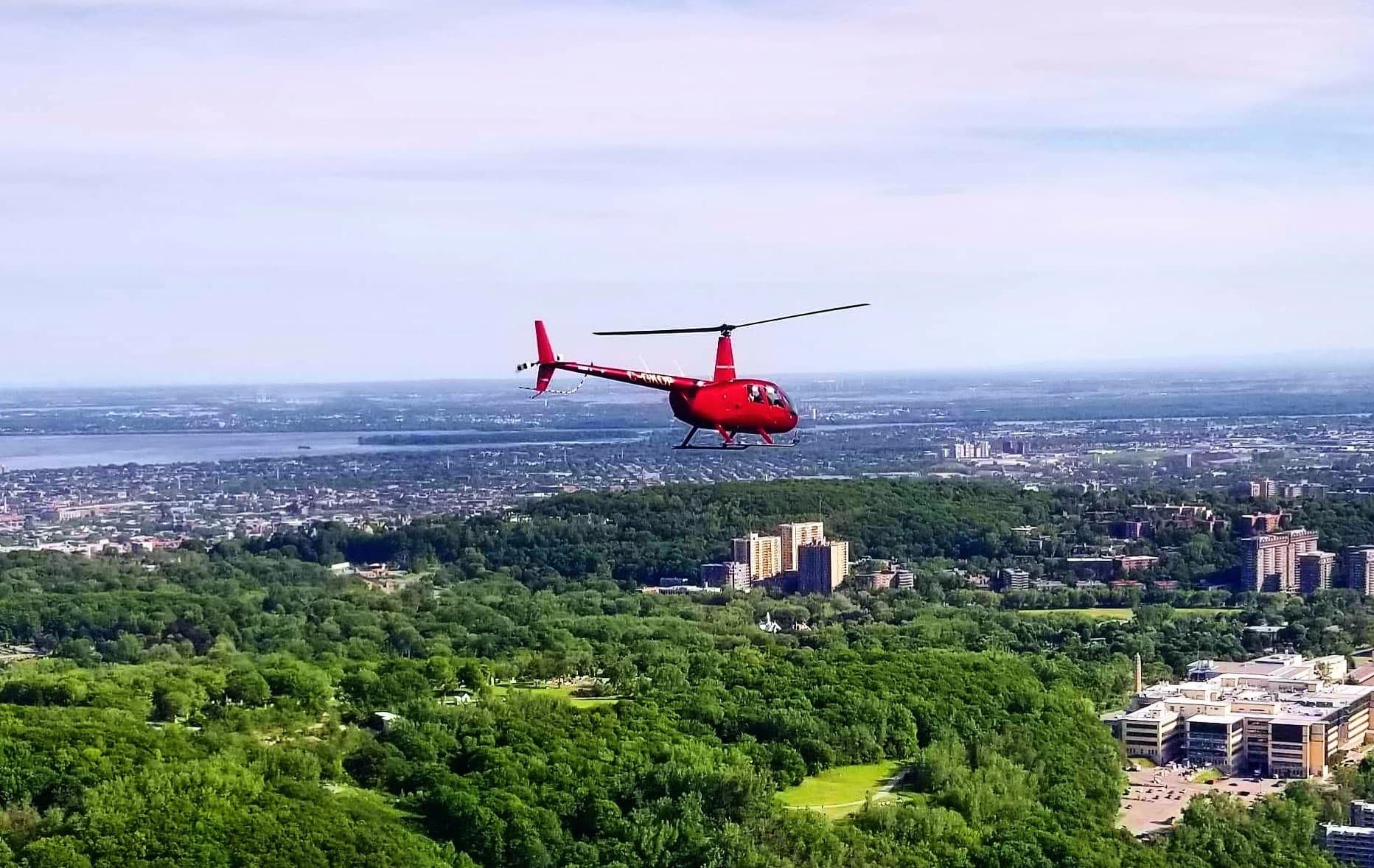 Red helicopter cruising above green forests and suburban landscape