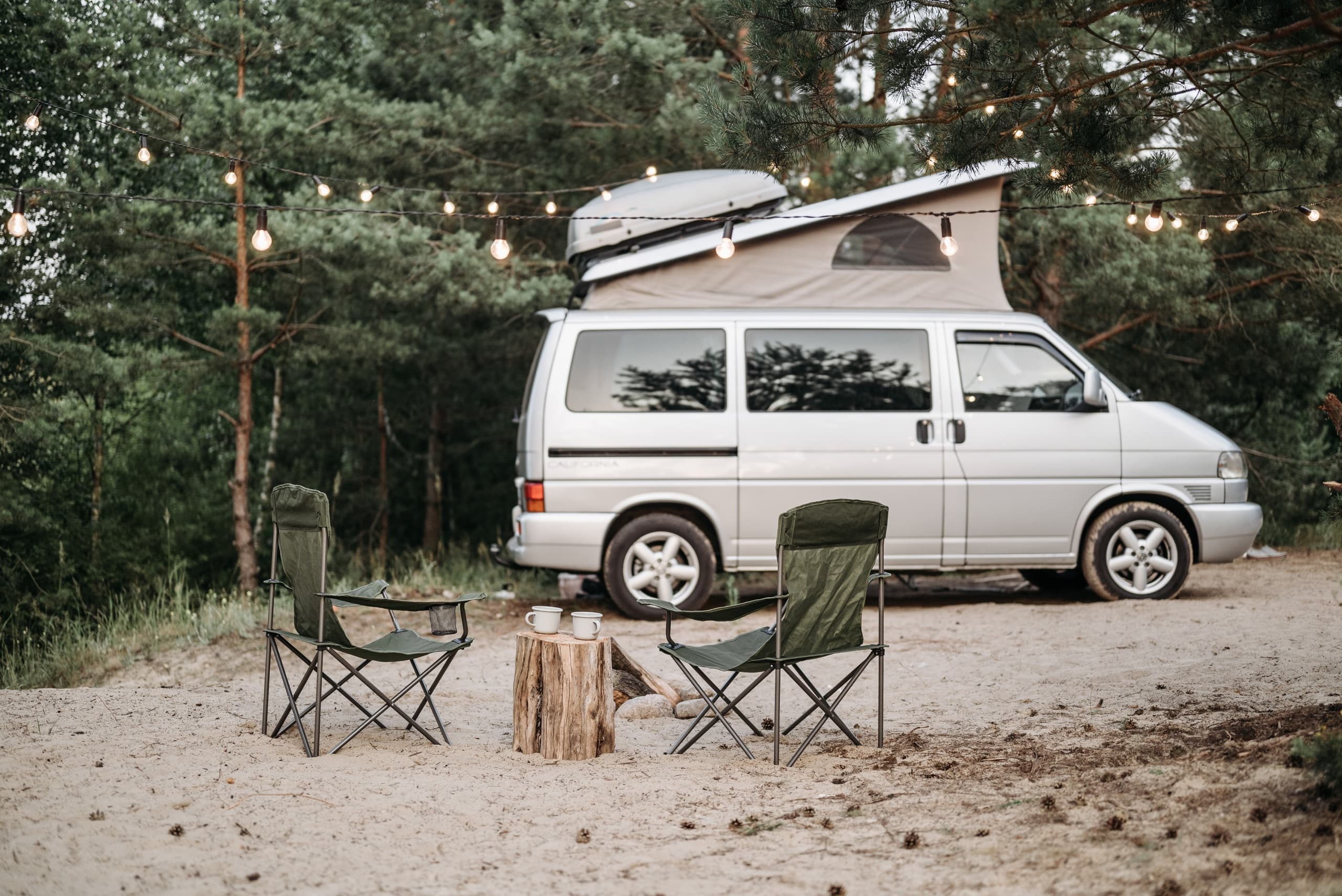 Camper van campsite with chairs in pine forest at dusk