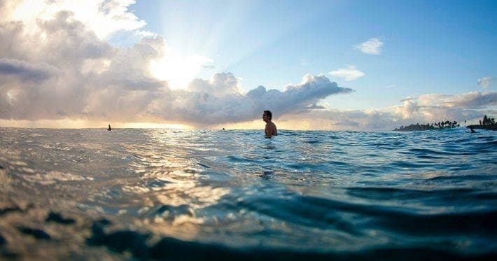 Surfer bobbing in ocean at sunrise, shimmering open water