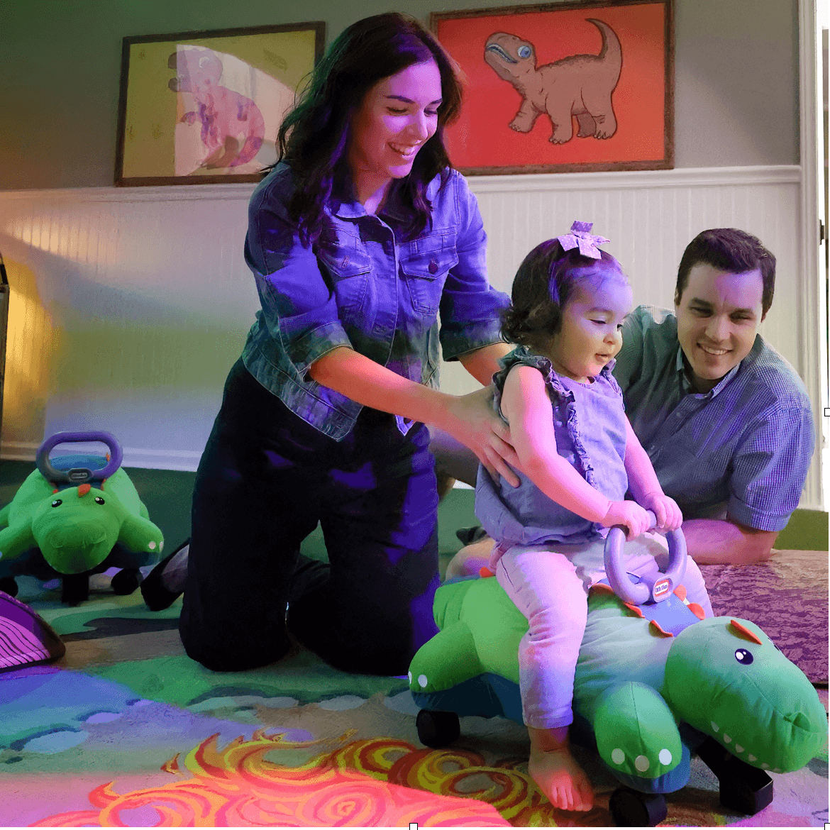 Parents help toddler ride toy dinosaur in colorful indoor playroom