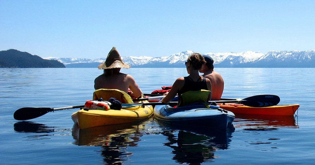 Three kayakers paddling on clear alpine lake with snowy peaks